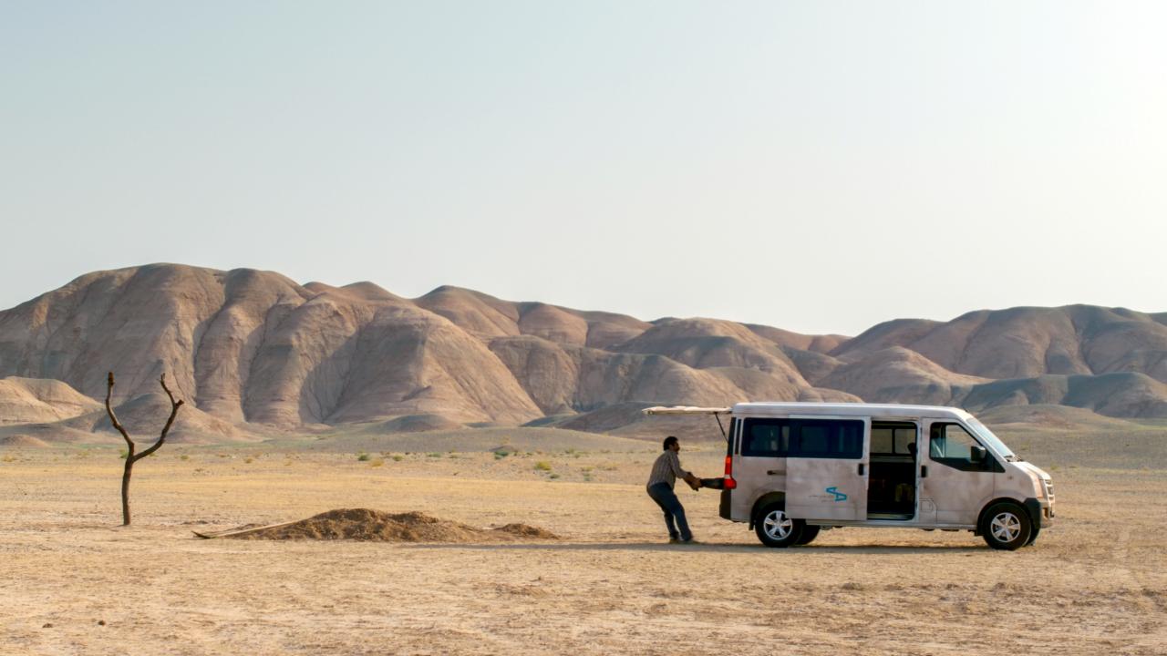 Filmstill aus dem Drama EIN EINFACHER UNFALL: Inmitten einer Wüstenlandschaft steht ein weißer Van. Ein Mann zieht an der hinteren Tür einen Menschen raus. Links neben ihm, ist ein Sandhaufen zu erkennen.