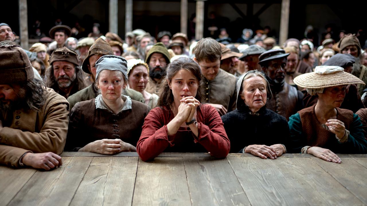 Filmstill aus dem Drama HAMNET: Vor der Bühne des Londoner Globe-Theaters im 16. Jahrhundert steht ein sichtlich ergriffenes Publikum vor der Bühne. In der Mitte steht eine rot gekleidete Frau, die ihre Hände wie zum Gebet geschlossen hat.