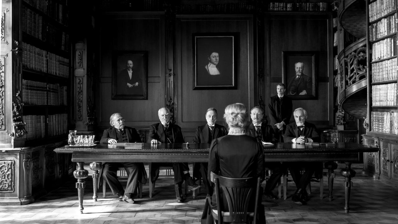 Filmstill aus SILENT FRIEND: Mit dem Rücken zum Betrachtenden sitzt eine junge Frau (gekleidet im Stil um 1900) vor einer Riege älterer Männer. Das Bild ist in Schwarz-Weiß.