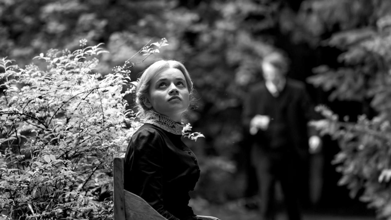 Schwarz-weißes Filmstill aus SILENT FRIEND: Eine junge Frau sitzt in einem Park auf einer Bank und schaut nach vorne gewandt leicht nach oben. Sie ist im Stil um 1900 gekleidet.