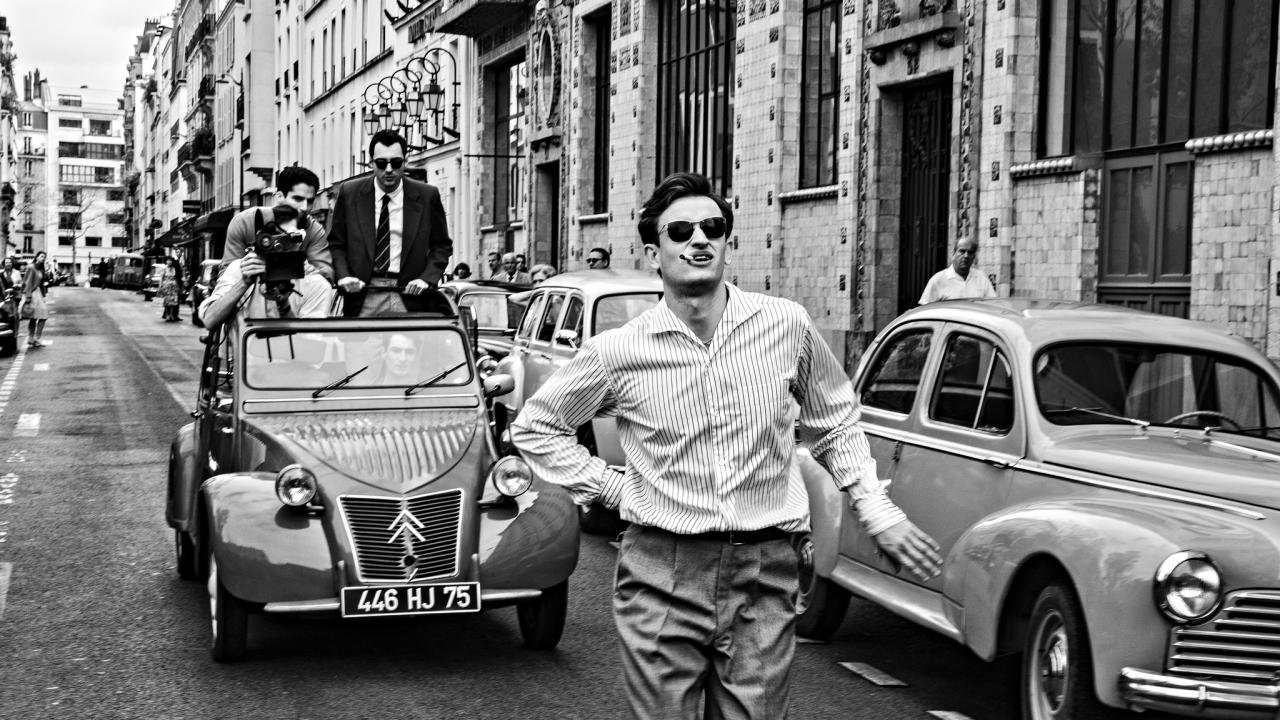 Filmstill aus NOUVELLE VAGUE: In der Schwarzweißaufnahme läuft ein junger Mann, der eine Sonnenbrille trägt und eine Zigarette raucht, in einer großstädtischen Seitenstraße, auf die Kamera zu. Direkt hinter ihm fährt ein Citroën 2CV,  durch dessen Faltdach drei Männer auf den Laufenden schauen. Einer hat eine Kamera auf ihn gerichtet.