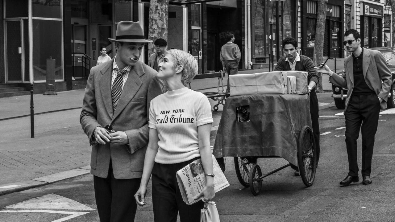 Still aus dem Biopic NOUVELLE VAGUE: Ein mit Sakko und Hut gekleideter Mann mit Zigette im Mund und eine Frau mit blonder Kurzhaarfrisur und im hellen T-Shirt mit der Aufschrift "New York Herold Tribune" gehen einander zugewandt auf einer Geschäftsstraße entlang. Ihnen folgen zwei Männer mit einem auffälligen großen Handwagen.