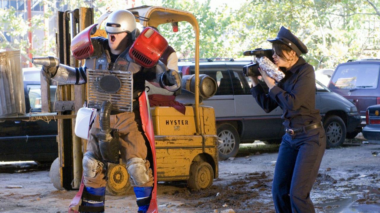 Filmstill aus der Komödie ABGEDREHT: Ein Mann, verkleidet als Titelfigur aus dem Sci-Fi-Film ROBOCOP, wird von einer Frau in Polizeikostüm mit einer kleinen Filmkamera aufgenommen.