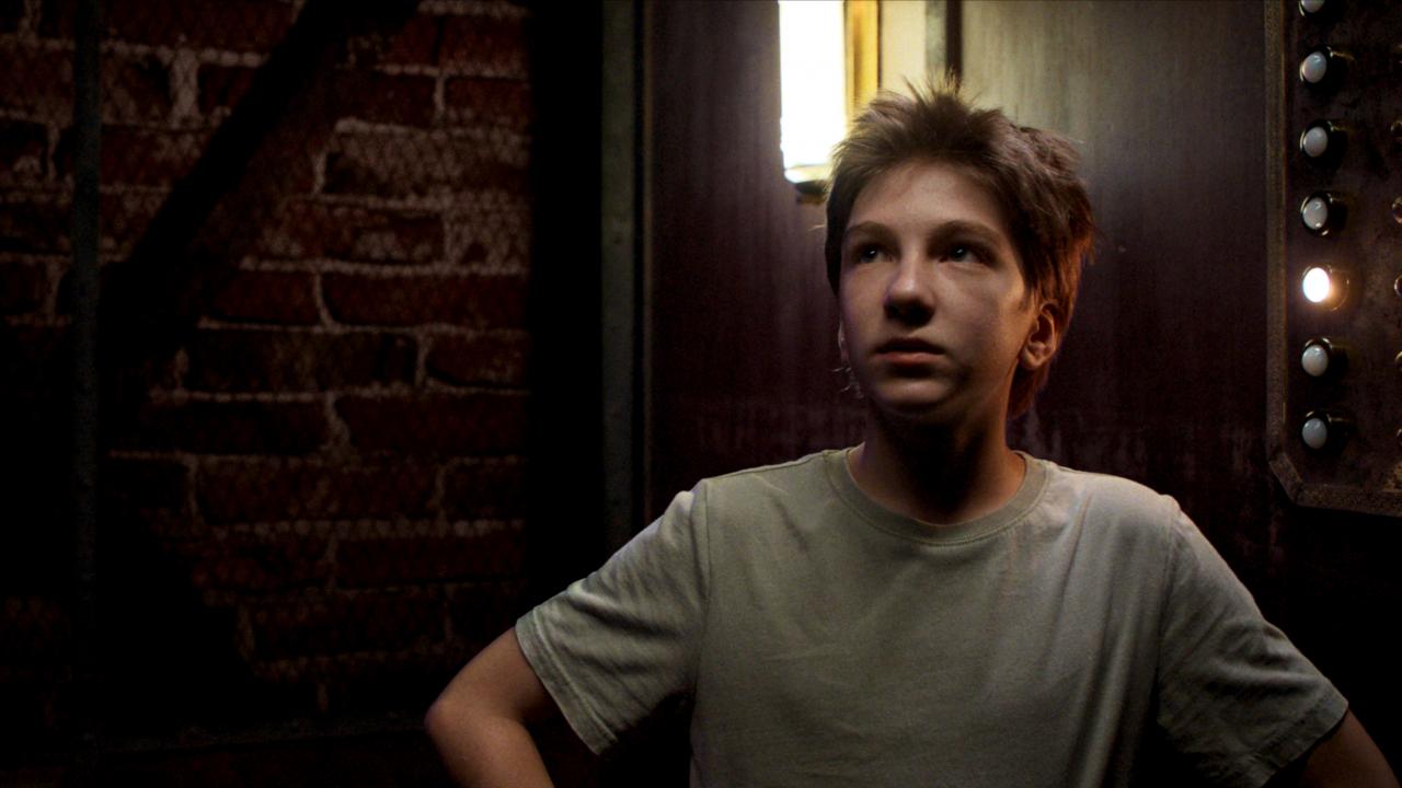 Still aus dem Kinderspielfilm DAS GEHEIME STOCKWERK: Ein etwa 12-jähriger Junge in T-Shirt steht in einem altmodischen, schwach beleuchteten Fahrstuhl. Hinter ihm leuchtet der Knopf der dritten Etage.