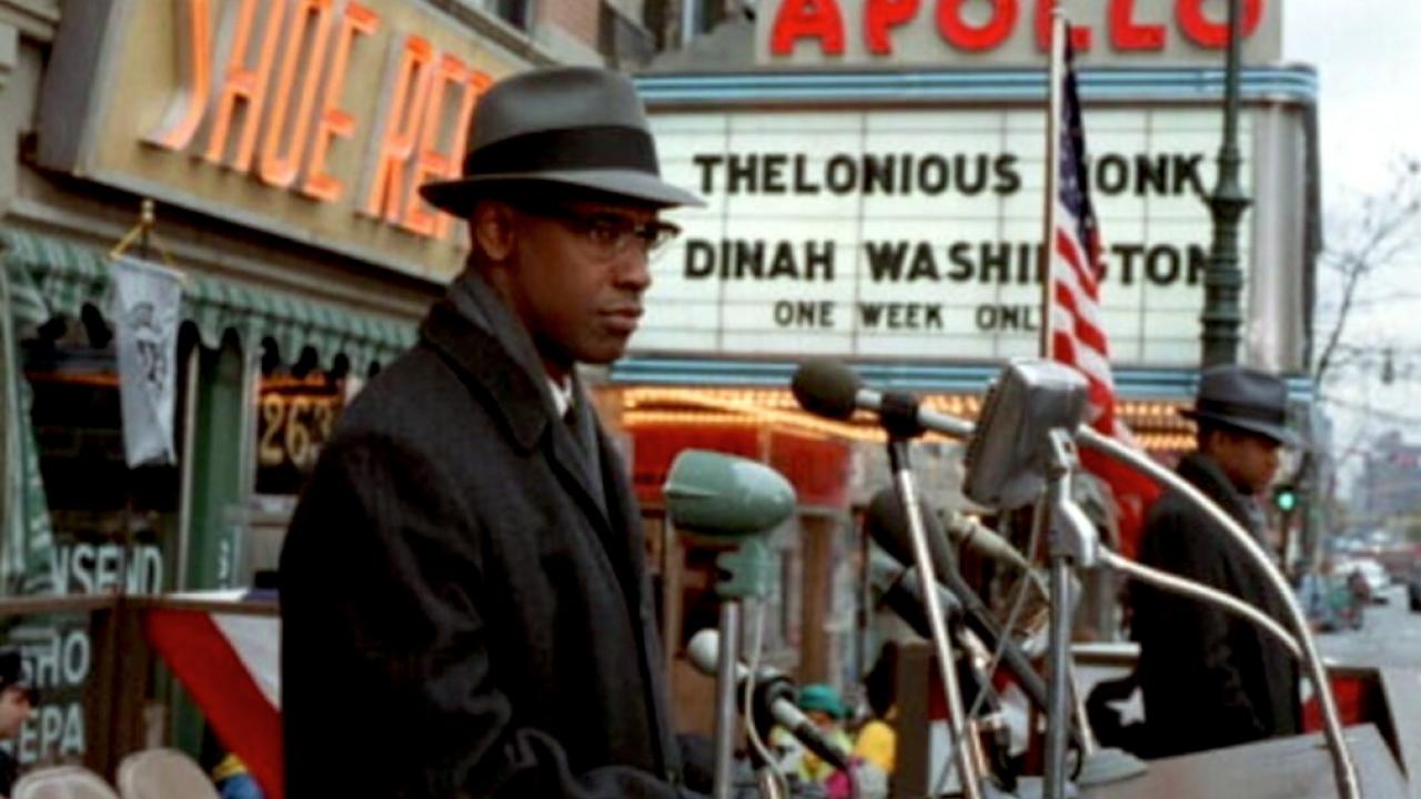 Filmstill aus MALCOLM X: Ein Mann mit Brille, Mantel und Hut steht auf einer Geschäftsstraße an einem Pult mit Mikrophonen.
