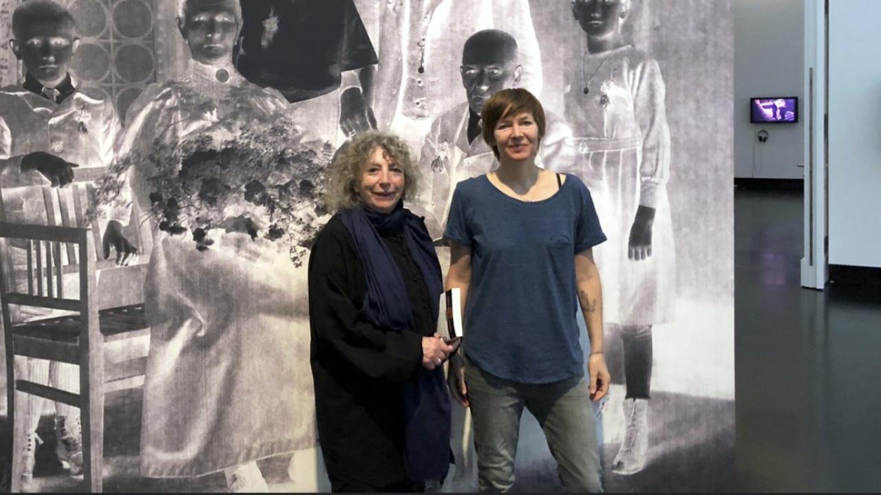 Amerikanische Aufnahme der Kamerafrau Elfi Mikesch und der Regisseurin Kerstin Polte, die in einer Ausstellung vor einer großen Schwarzweiß-Fotografie stehen.