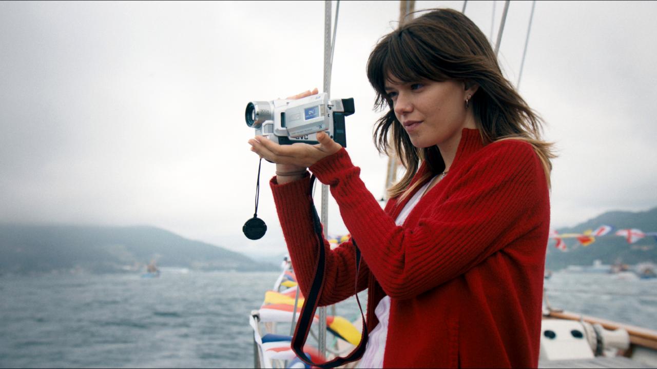 Filmstill aus dem Drama ROMERÍA – DAS TAGEBUCH MEINER MUTTER: Eine junge Frau in roter Strickjacke steht auf einer Segeljacht und filmt mit einer kleiner Videokamera. Der Himmel ist bewölkt.