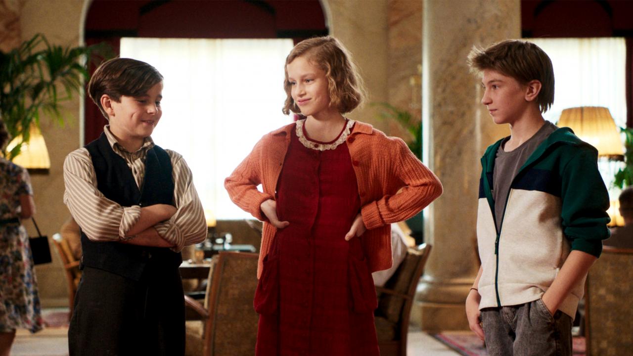 Still aus dem Kinderspielfilm DAS GEHEIME STOCKWERK: Zwei Jungen und ein Mädchen stehen nebeneinander in einer altmodischen Hotellobby. Während der eine Junge modern gekleidet ist, sind die anderen im Stil der späten 1930er-Jahre angezogen und frisiert.