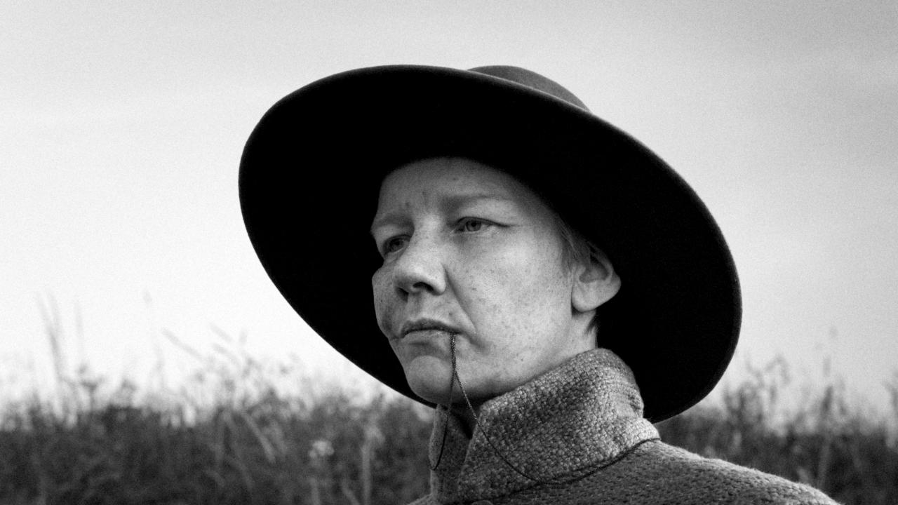 Still aus dem Drama ROSE: Schwarz-Weiß-Porträt einer Person, die androgyn wirkt. Sie trägt einen altmodischen, breitkrempigen Hut und hat den Anhänger einer Kette im Mund.