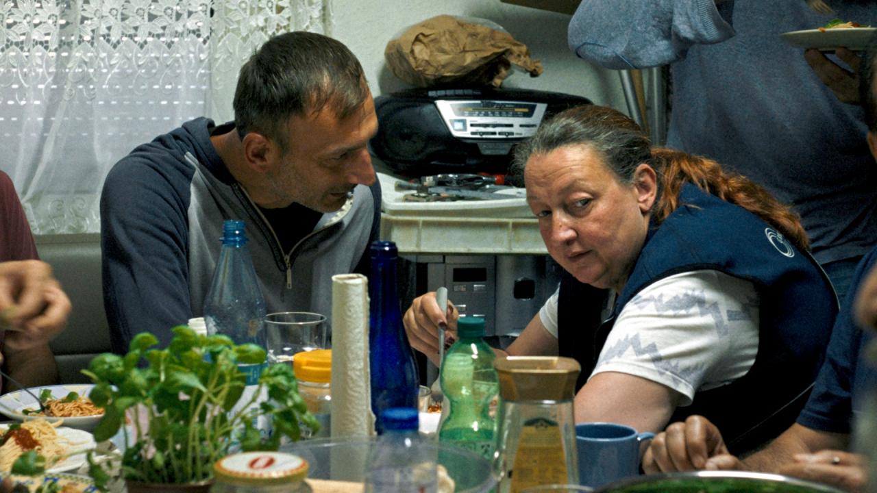 Still aus dem Drama ICH VERSTEHE IHREN UNMUT: Eine Frau, Ende 50, sitzt in einem Pausenraum an einem Tisch. Ein Mann redet auf sie ein.