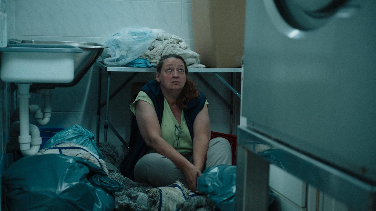 Still aus dem Film ICH VERSTEHE IHREN UNMUT: Halbtotale einer Frau die in einer Waschküche zwischen Wischlappen und Müllsäcken auf dem Boden sitzt und nach oben blickt.