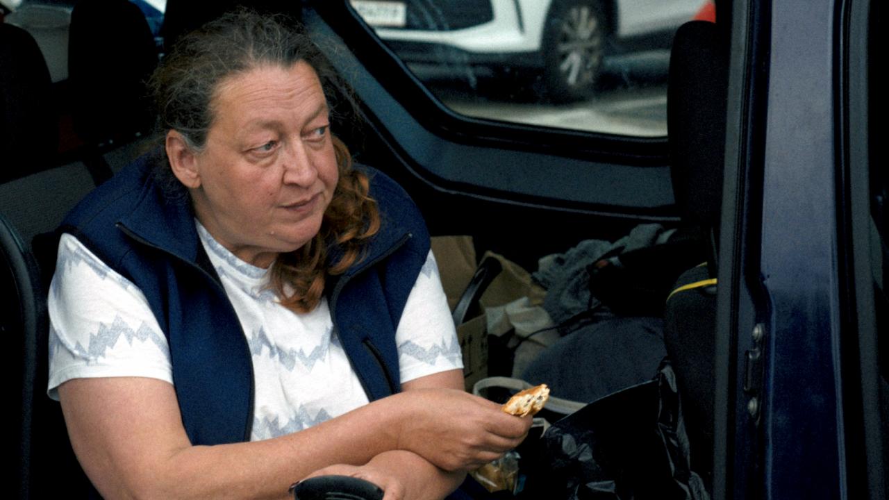 Still aus dem Film ICH VERSTEHE IHREN UNMUT: Eine Frau sitzt bei geöffneter Tür in einem Auto. In der einen Hand hält sie einen Becher, in der anderen ein belegtes Brot.