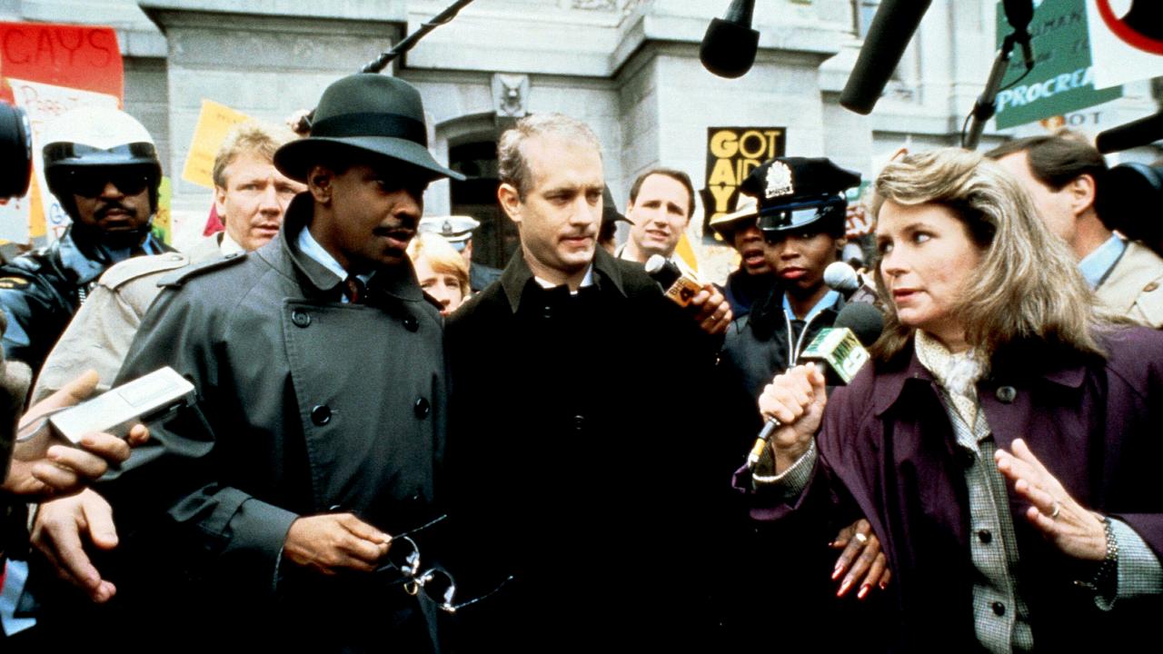 Still aus dem Drama PHILADELPHIA: Ein schwarzer und ein weißer Mann stehen in einer Menschenmasse vor einem Gebäude. Eine Reporterin hält ein Mikrofon in der Hand und stellt ihnen eine Frage.