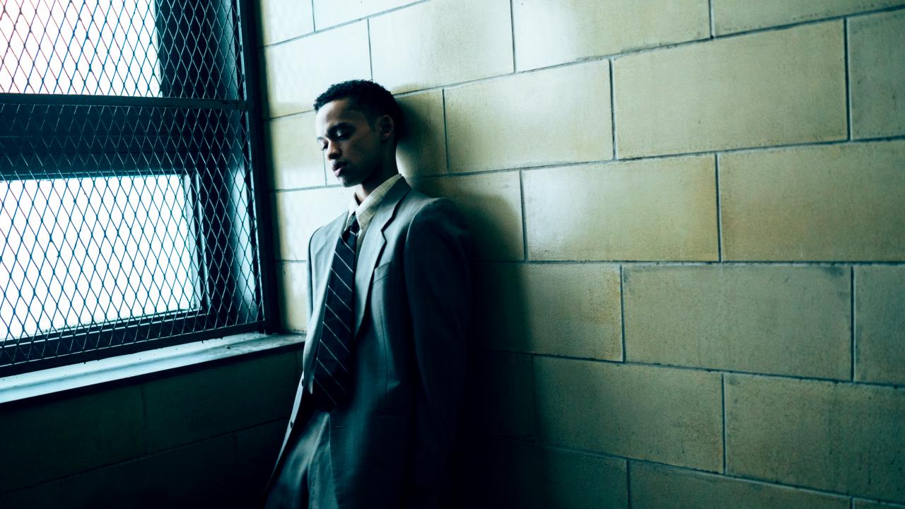 Still aus der Serie WHEN THEY SEE US: Ein junger Schwarzer im Anzu steht an an einem vergitterten Fenster und lehnt sich an eine gekachelte Wand an.