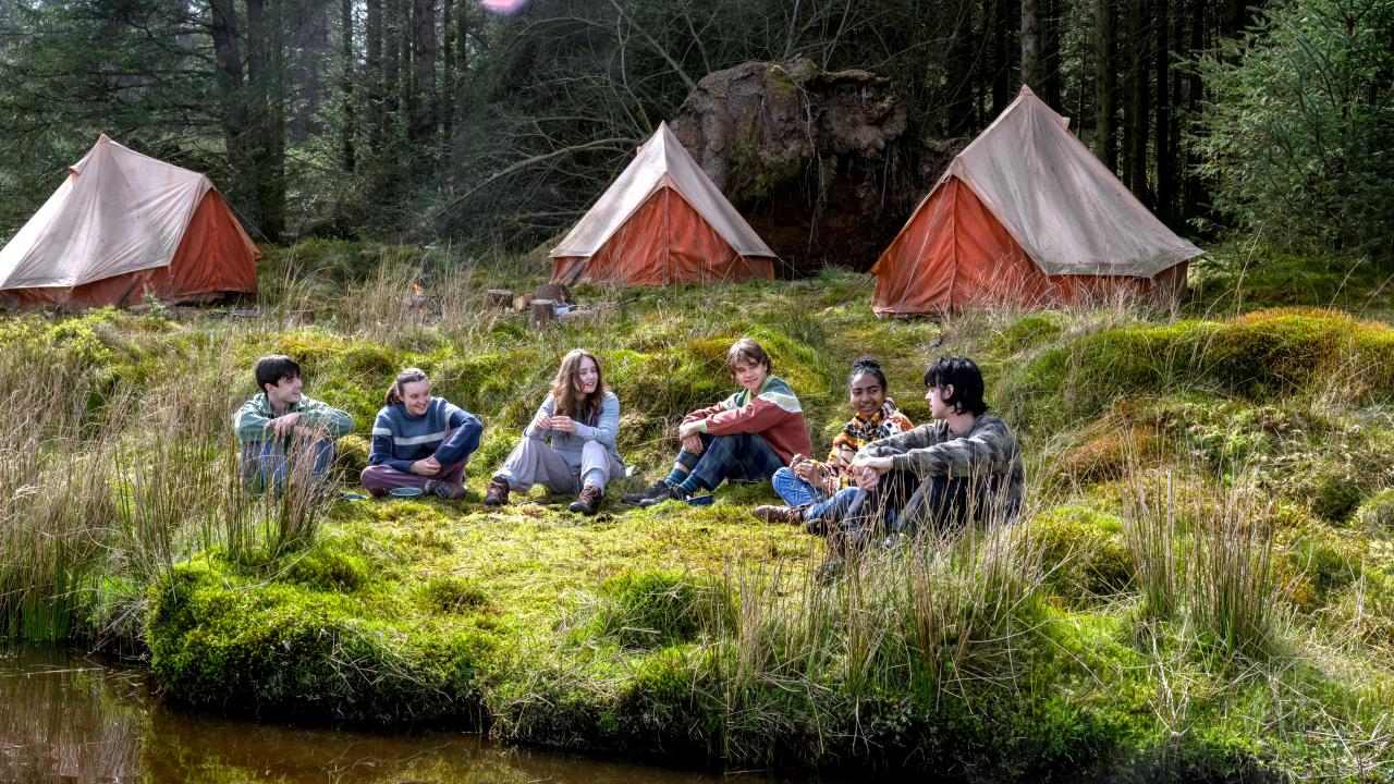 Still aus dem Drama SUNNY DANCER: An einem Seeufer sitzt eine Gruppe Jugendlicher im Gras. Im Hintergrund sind drei Zelte zu erkennen.