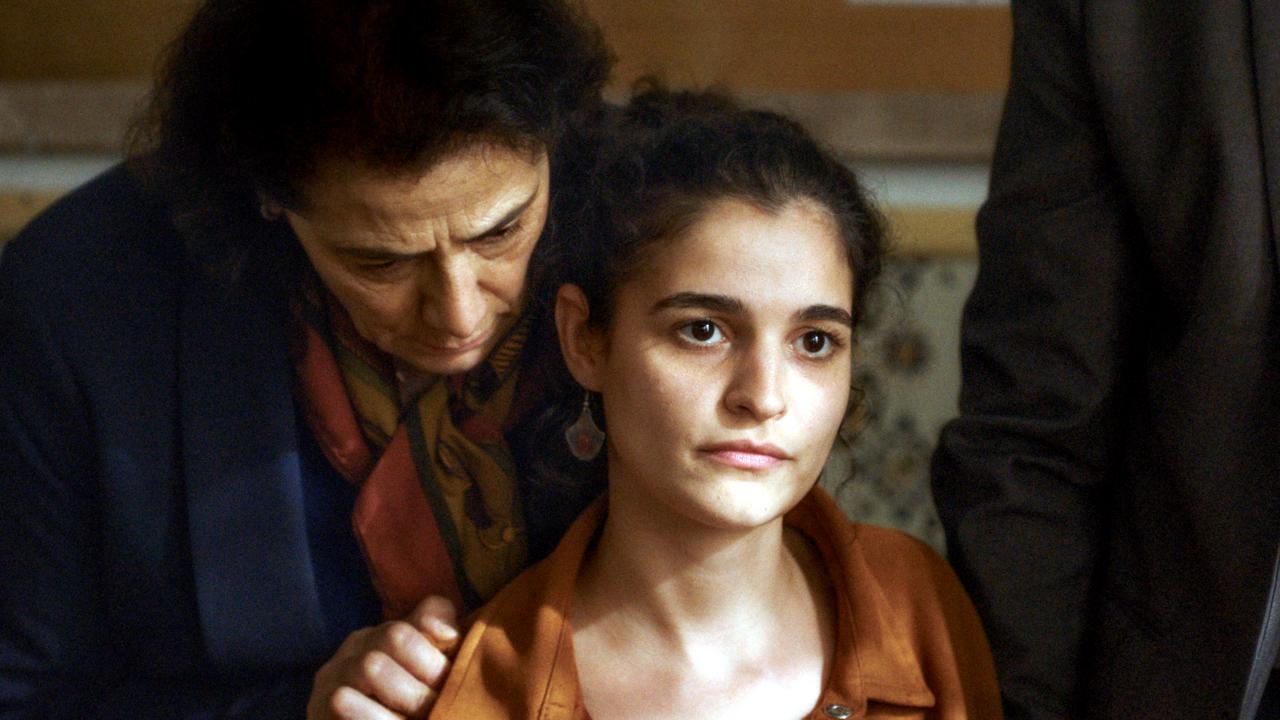 Still aus dem Drama MIT LEISER STIMME: Nahaufnahme einer jungen Frau. Hinter ihr steht eine ältere Fraum die sich zu ihr herunterbeugt.