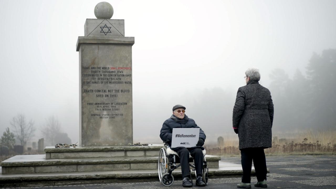 Still aus dem Dokumentarfilm ALBRECHT WEINBERG – ES IST IMMER IN MEINEM KOPF: In der Gedenkstätte Bergen Belsen sitzt Albrecht Weinberg im Rollstuhl vor einem Gedenkstein. Er hält ein Schild mit der Aufschrift "#We remember"  vor sich. Vor ihm steht eine Frau, mit dem Rücken zur Kamera. Es ist neblig, die beiden sind warm angezogen.