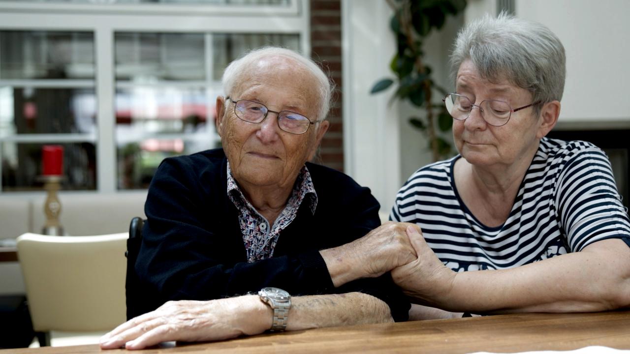Still aus dem Dokumentarfilm ALBRECHT WEINBERG – ES IST IMMER IN MEINEM KOPF: Albrecht Weinberg und Gerda Dänekas sitzen nebeneinander an einem Tisch. Sie hält seine Hand.