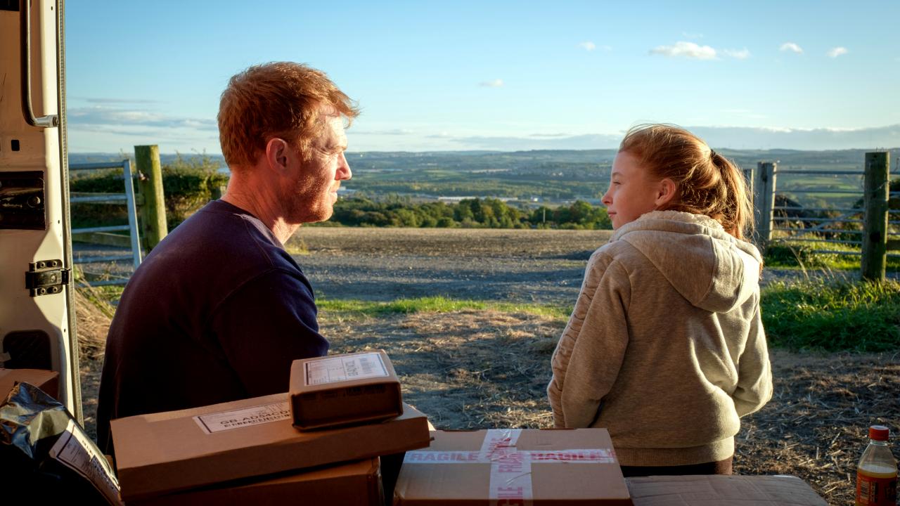 Still aus SORRY, WE MISSED YOZ: Ein Vater und seine Tochter sitzen auf der Ladefläche eine Lieferwagens und schauen sich an. Man sieht eine weite Landschaft.