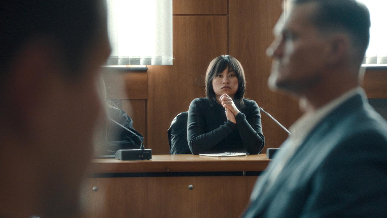 Still aus dem Thriller STAATSSCHUTZ: Eine junge, deutsche-koreanische Frau sitzt in einem holzgetäftelten Gerichtssaal. Sie ist frontal zu sehen und hat die Hände gefaltet. Im Vordergrund ist ein Mann im grauen Anzug unscharf im Profil zu sehen.