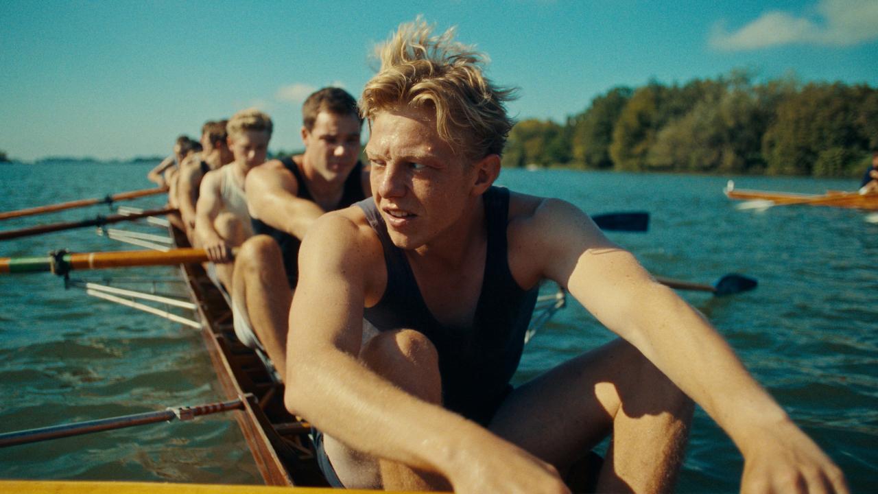 Szenenbild zum Film ADAMS ACHT: Auf einem See sitzt eine Gruppe junger Männer in Unterhemden und kurzen Hosenin einem Sportruderboot. Rechts im Hintergrund ist unscharf ein weiteres Ruderboot zu erkennen.