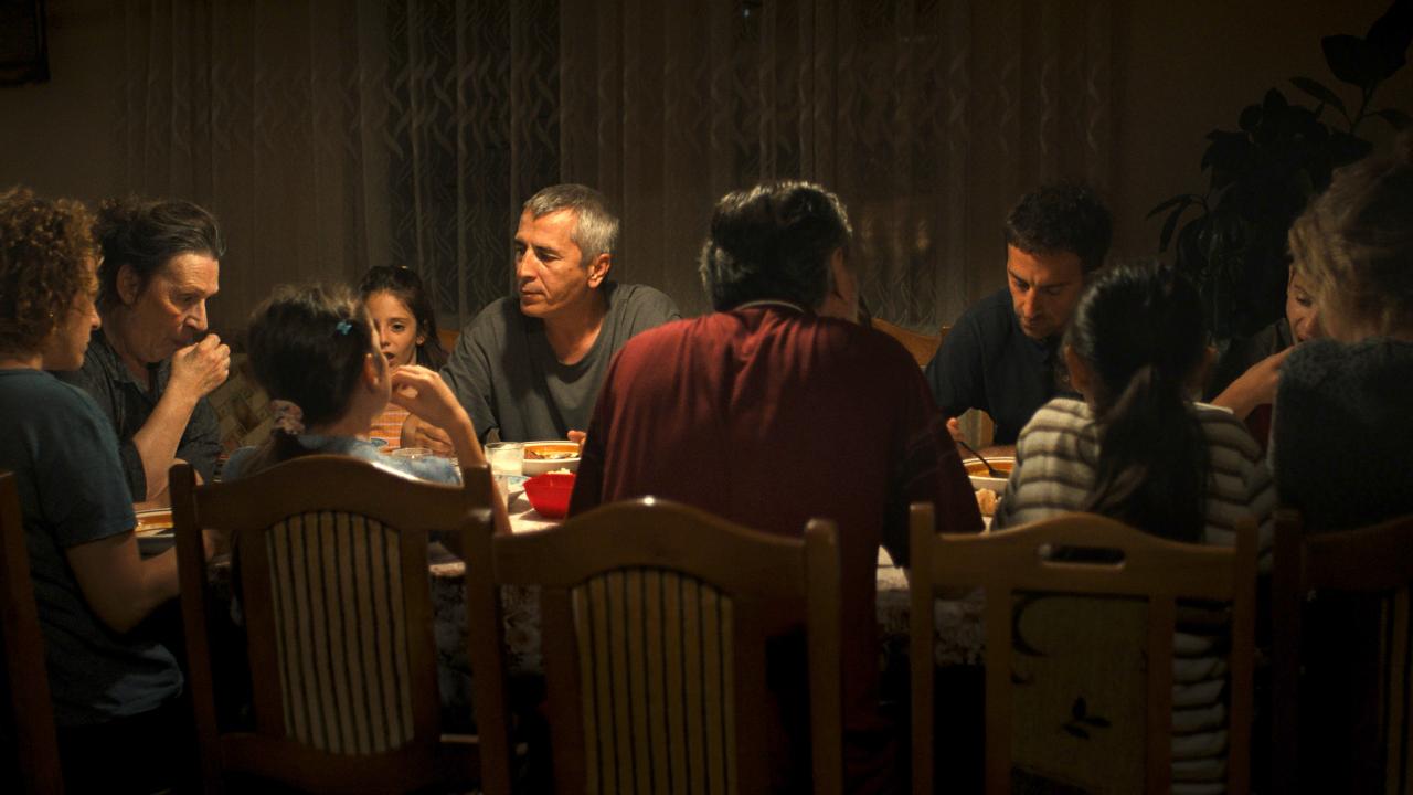 Still aus dem Drama VON SCHAM UND GELD: An einem großen Esstisch sitzen Männer und Frauen und Kinder in einem eher dunklen Raum. Auf einem Mann mit kurzen grauen Haaren fällt Licht von oben. 