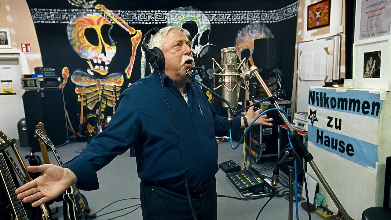 Still aus dem Dokumentarfilm BIERMANN: Wolf Biermann im Studio der Toten Hosen bei den Aufnahmen zu seinem Lied „Ermutigung“, das er gemeinsam mit der Band aufgenommen hat. Er Steht mit ausgestreckten Armen singend vor einem Mikrofon.