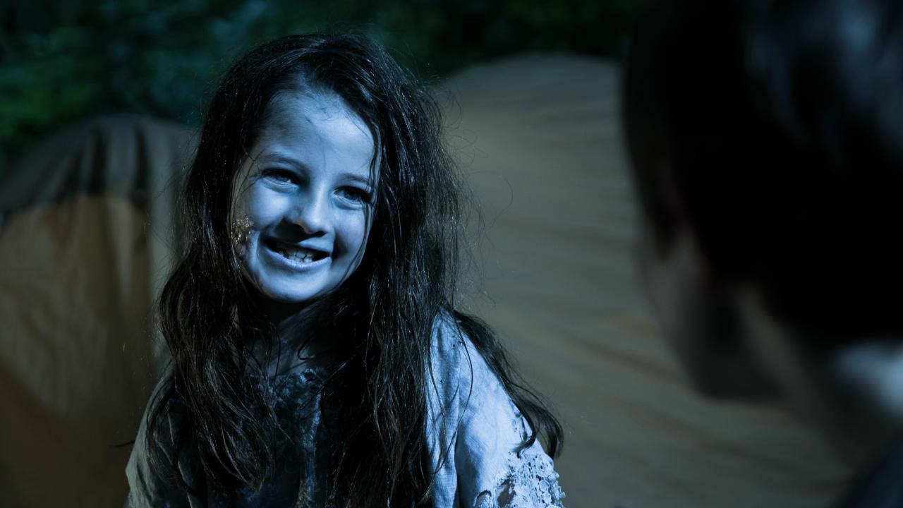 Still aus dem Kurzfilm ZOMBIERELLA: Ein lächelndes Mädchen mit ungesund fahler Gesichtsfarbe, zerzaustem Haar und zerschlissener Kleidung fletscht lächelnd die Zähne während es eine unscharf am rechten Bildrand zu erahnende Person anschaut.