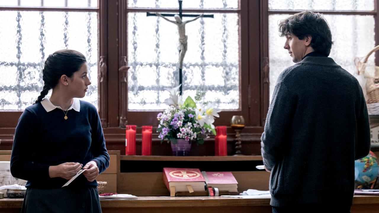 Still zum Drama GLAUBE: Ein Mädchen und ein Junge, beide sind Teenager, stehen vor einem vergitterten Fenster. Sie schauen sich an. Auf dem Fesnterbrett liegt eine Bibel, es sind Kerzen und ein Kruzifix zu sehen. 
