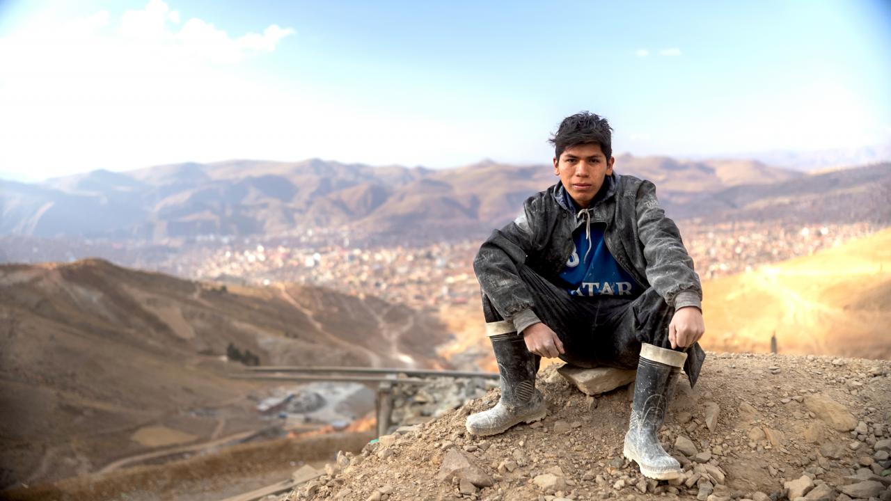 Still aus dem Dokumentarfilm MATERIA PRIMA: Ein junger Mann sitzt auf dem Boden und schaut direkt in die Kamera. Im Hintergrund ist eine karge Hügellandschaft in Bolivien zu erkennen.