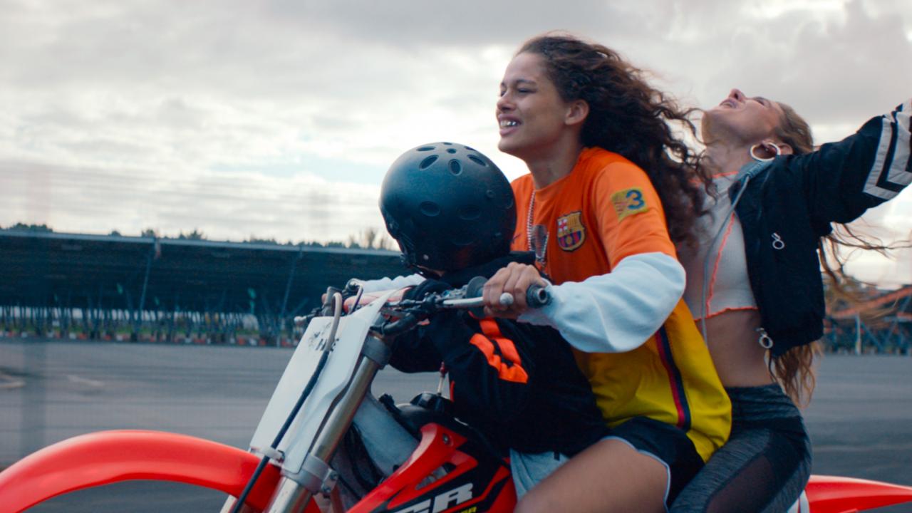 Still aus dem Drama RODEO: Zwei junge Frauen und ein Kind sitzen auf einem Motorrad. Die Frau, die hinter der Fahrerin sitzt, reißt ihre Arme in die Höhe. Nur das kleine Kind trägt einen Sturzhelm.