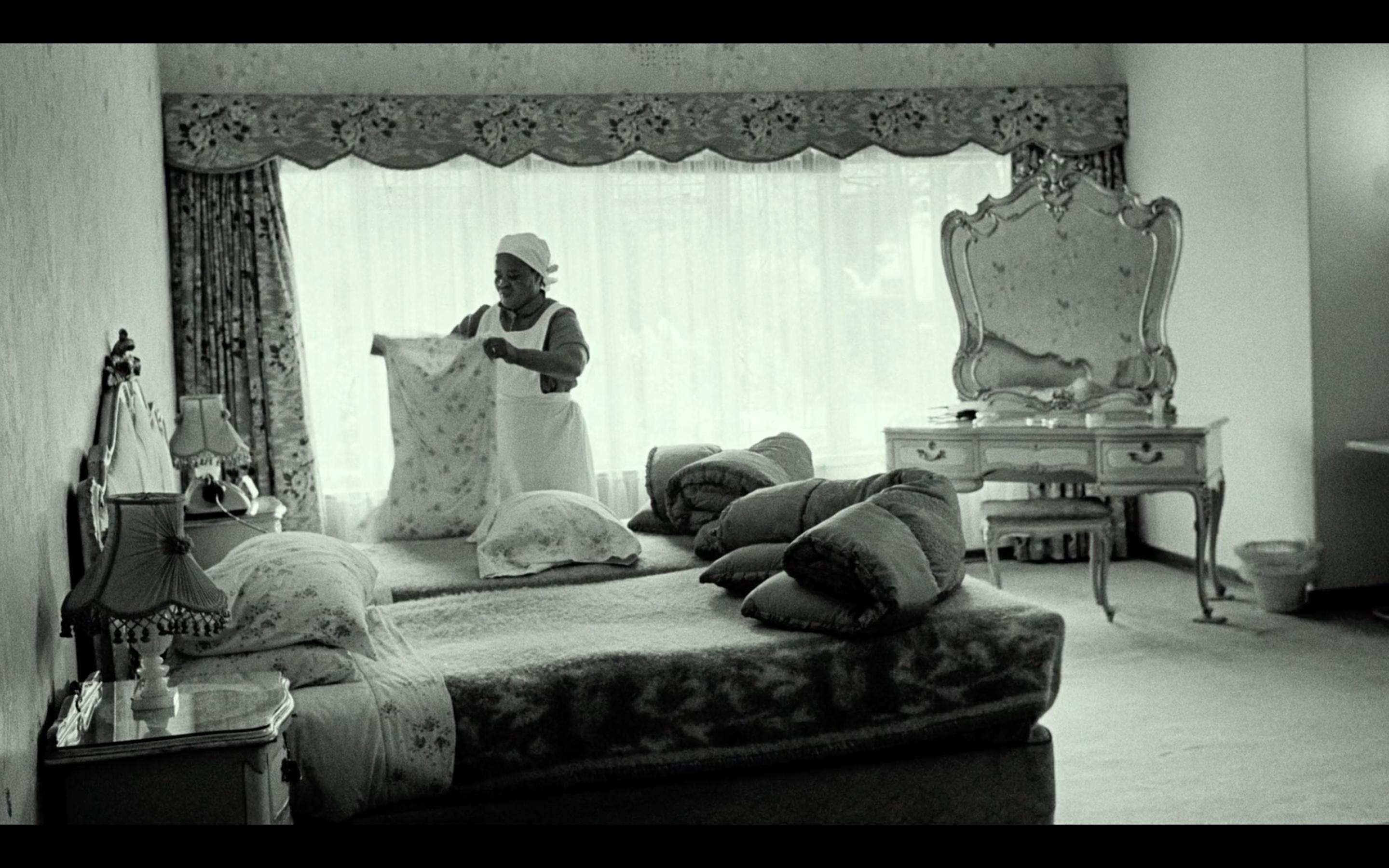 Schwarz-Weiß-Foto: In einem elegant eingerichteten Schlafzimmer richtet eine Haushälterin afrikanischer Herkunft das Bett, während weiches Licht durch ein großes Fenster mit schweren Vorhängen fällt.