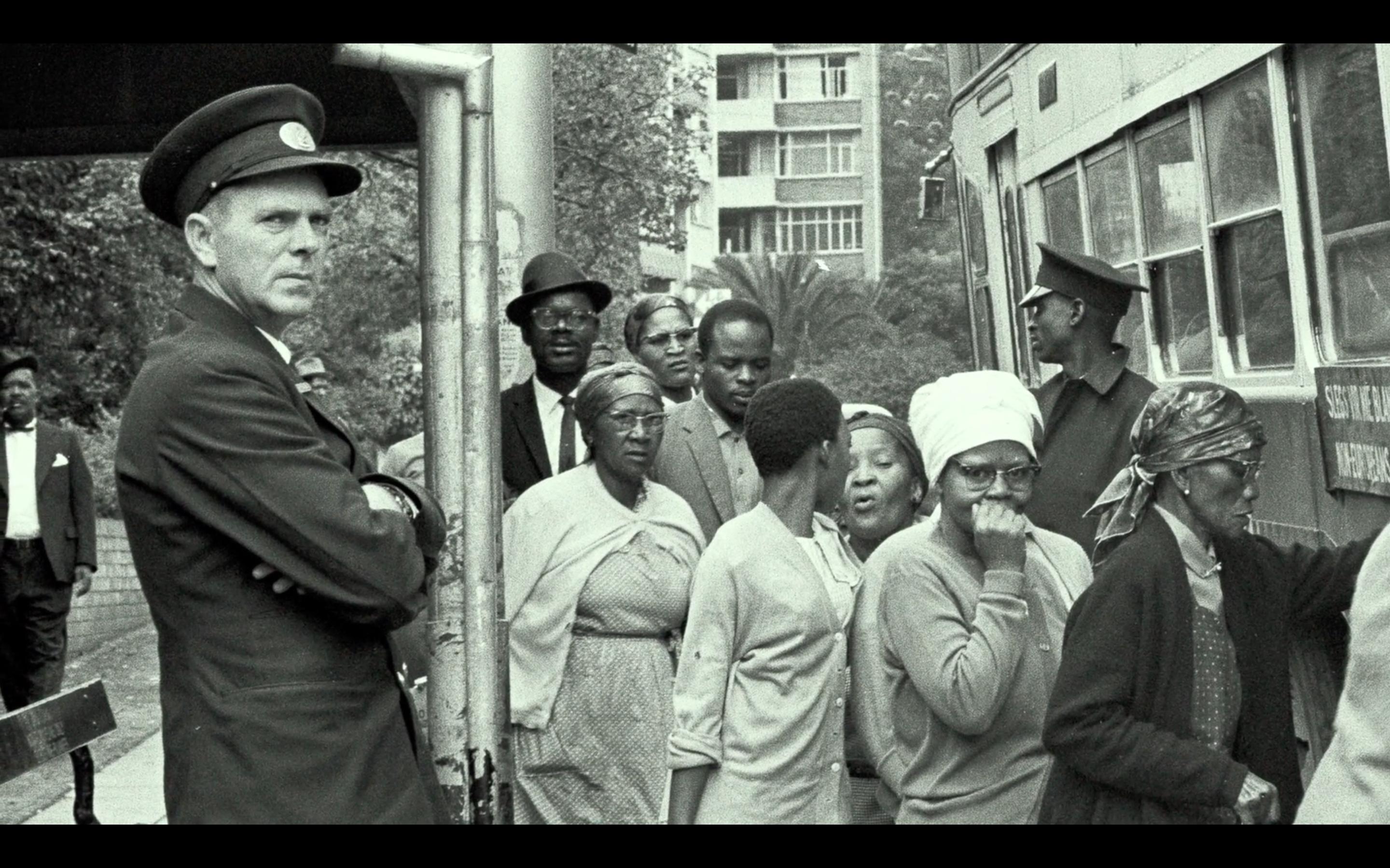 Links in dem Schwarz-Weiß-Foto ist ein uniformierter weiß gelesener Mann in Uniform zu sehen. Daneben befinden sich mehrere schwarz gelesene Personen, die ihn passieren.