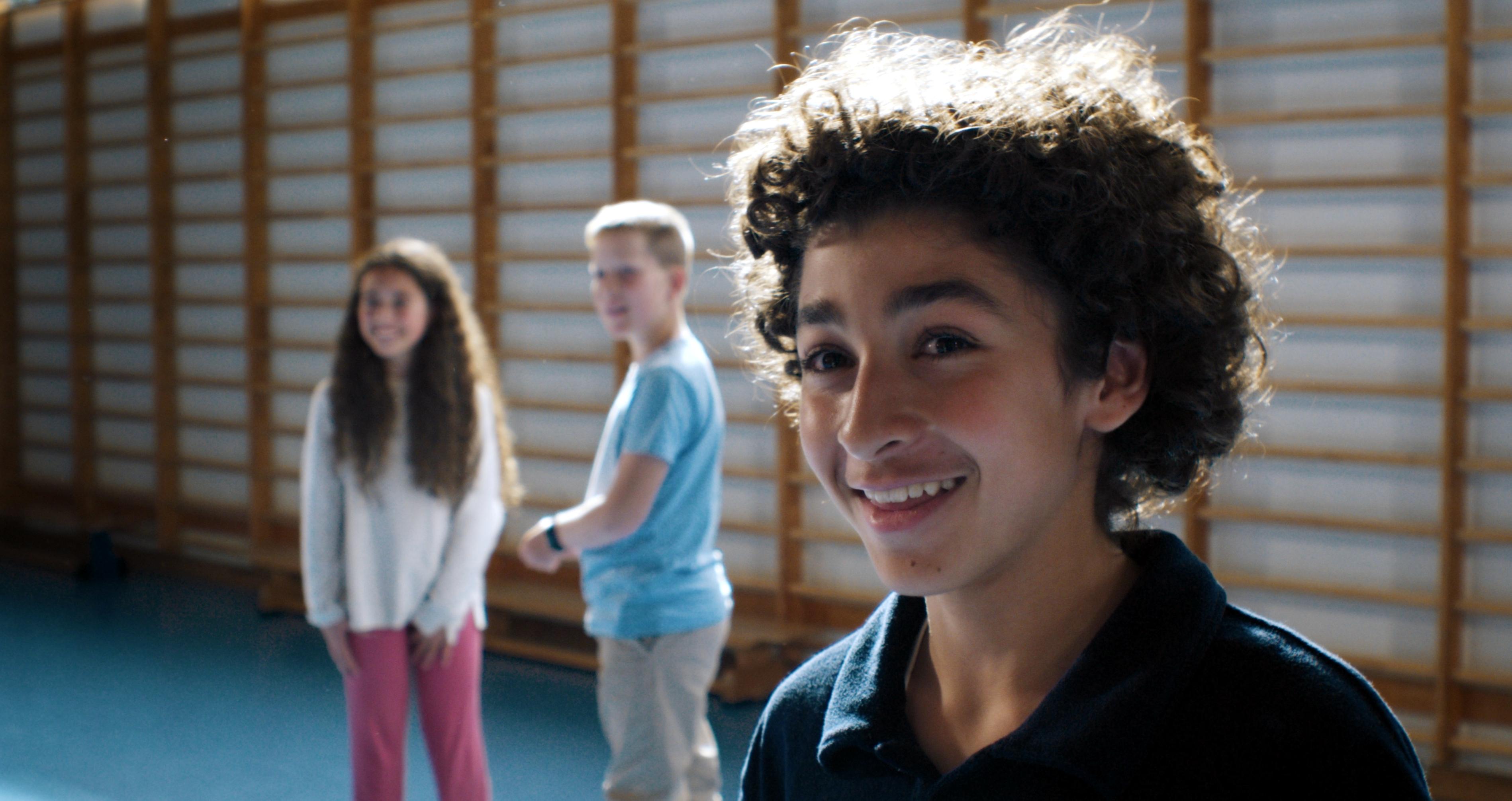 Still aus dem Kinderfilm LARS IST LOL: Porträt eines lächelnden Jungen, der in einer Turnhalle steht. Hinter ihm sind zwei Kinder zu sehen.