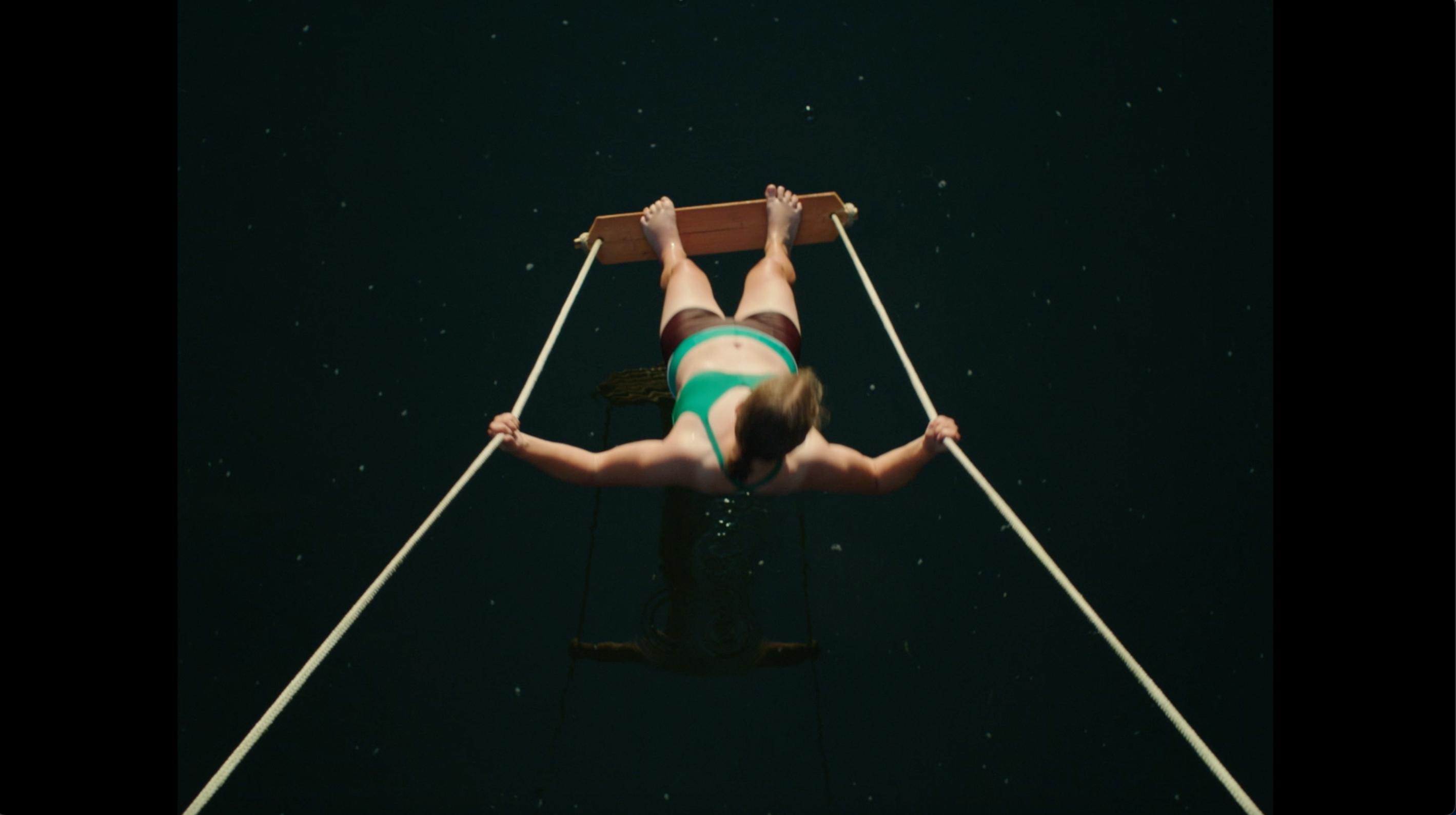 Screenshot aus dem Drama MILCH INS FEUER: Aus der Obersicht sieht man eine junge Frau im Bikini, die stehend auf einer Schaukel über Flusswasser schwingt.