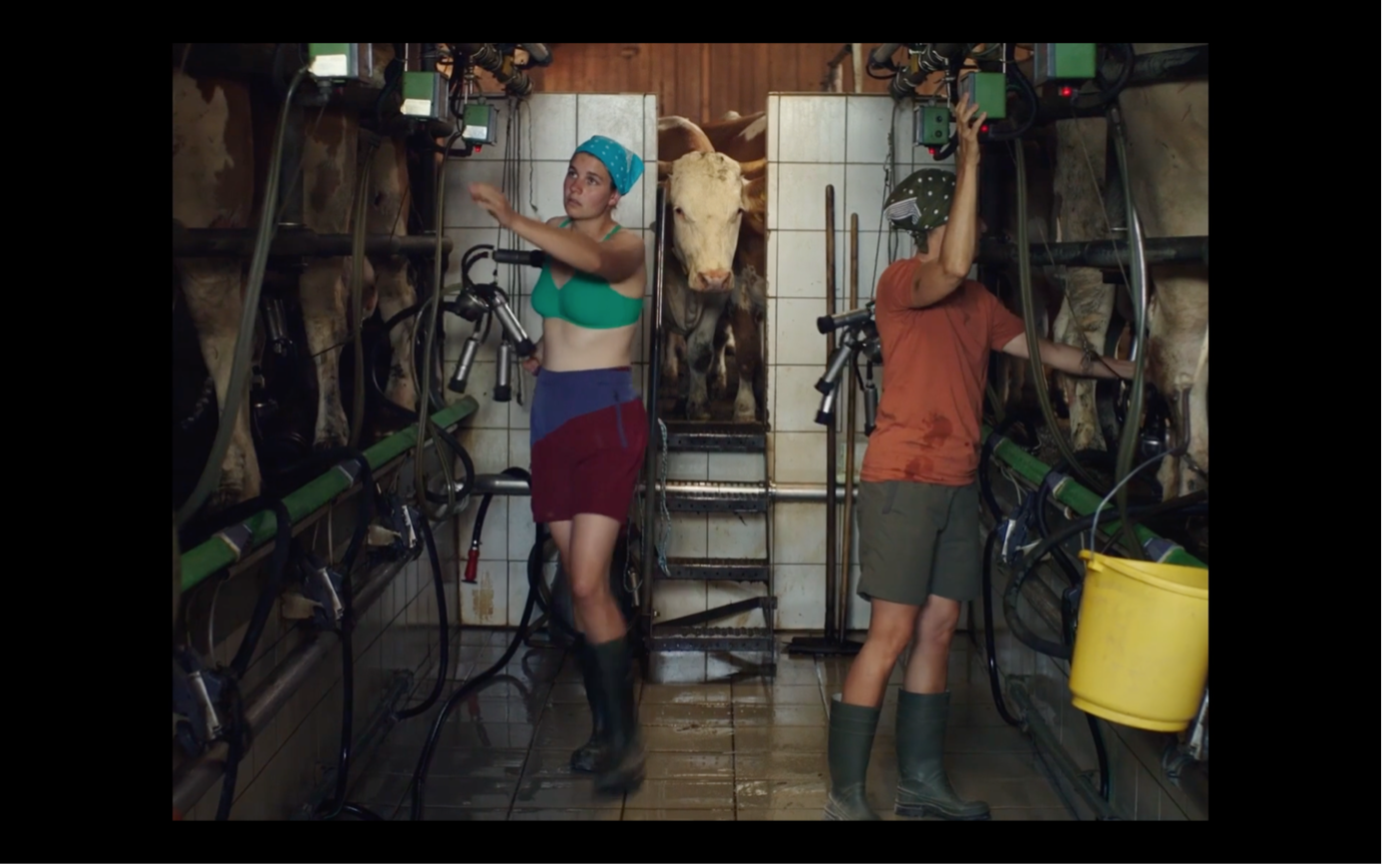 Screenshot aus MILCH INS FEUER: Katinka und ihre Mutter im Stall beim Melken. Beide tragen Kopftücher, Gummistiefel und kurze Hosen. Links und rechts befinden sich Kühe.