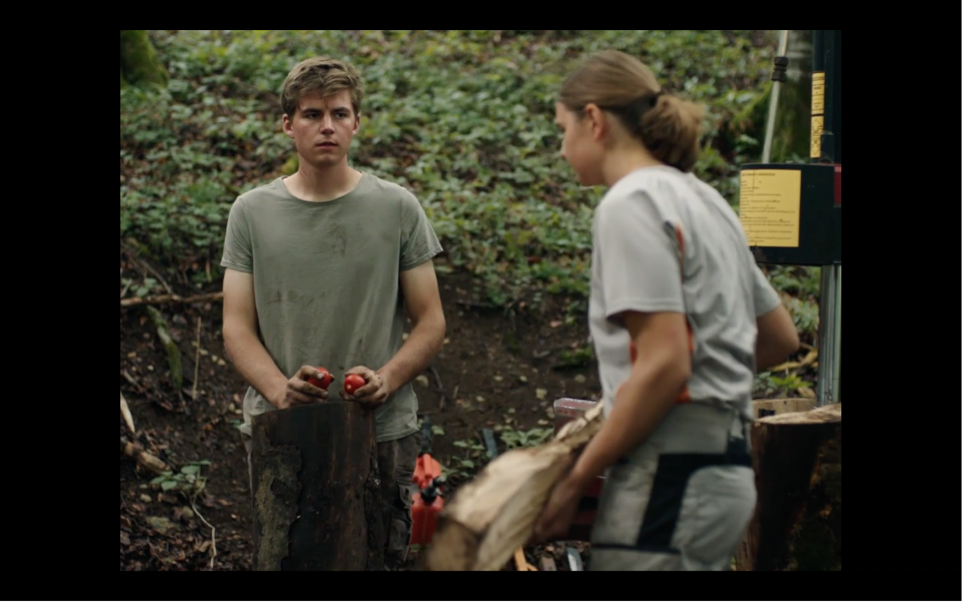 Screenshot aus MILCH INS FEUER: Katinka hält einen Holzstamm in der Hand und hört ihrem Bruder zu. Beide befinden sich im Garten.
