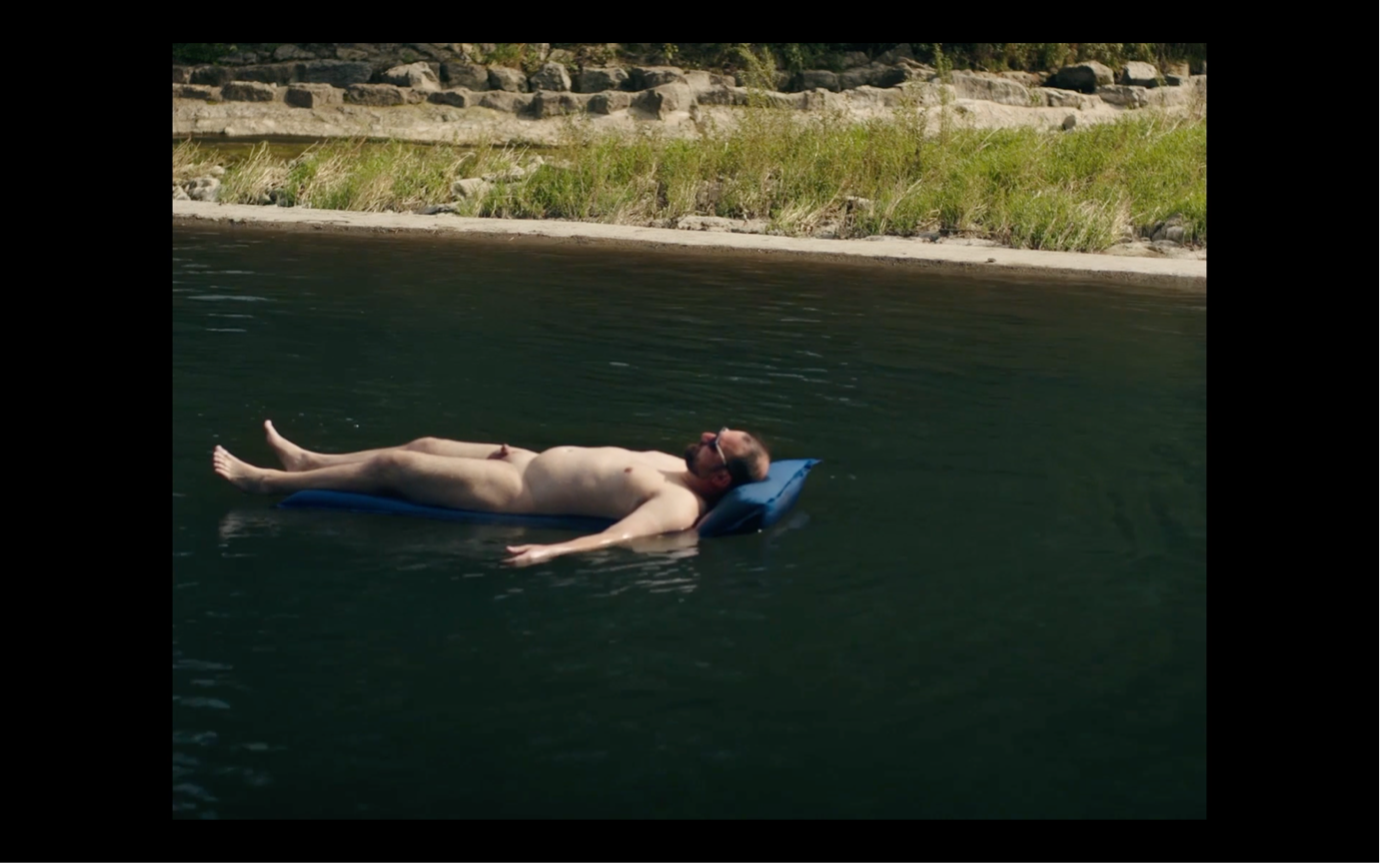 Screenshot aus MILCH INS FEUER: Ein nackter Mann treibt auf dem Rücken liegend auf einer Luftmatratze auf dem Fluss.