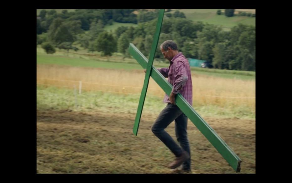 Screenshot aus MILCH INS FEUER: Ein Bauer geht auf einer Wiese und trägt ein etwa zwei Meter großes grünes Kreuz.