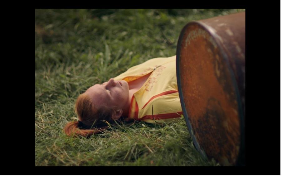 Screenshot aus MILCH INS FEUER: Die Freundin von Katinka liegt auf einer Wiese vor einem Faß.