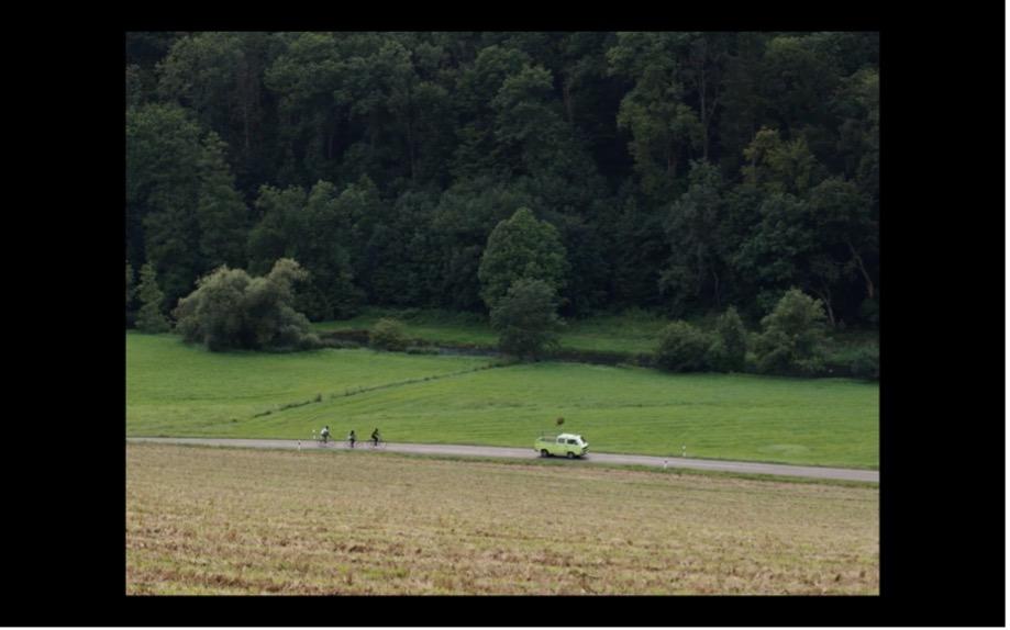 Screenshot aus MILCH INS FEUER: Ein Auto fährt auf einer Landstraße, dahinter drei Fahrradfahrer.