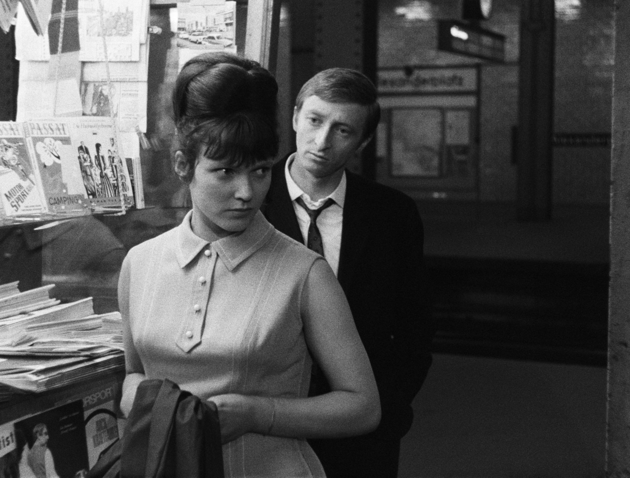 Filmstill aus dem DEFA-Film JAHRGANG 45: Eine junge Frau steht vor einem Geschäft und blickt leicht nach hinten. Hinter ihr steht ein junger Mann, der sie fragend anschaut.