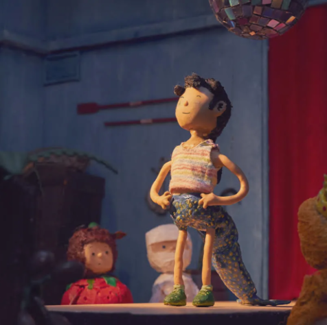 Eine kleine Stop-Motion-Figur steht selbstbewusst auf einer Bühne, die Hände in die Hüften gestemmt und den Kopf leicht erhoben. Umgeben von bunten Requisiten und warmem Licht wirkt die Szene wie ein liebevoll gestaltetes Puppentheater, mit einem schimmernden Diskokugel-Detail im Hintergrund.