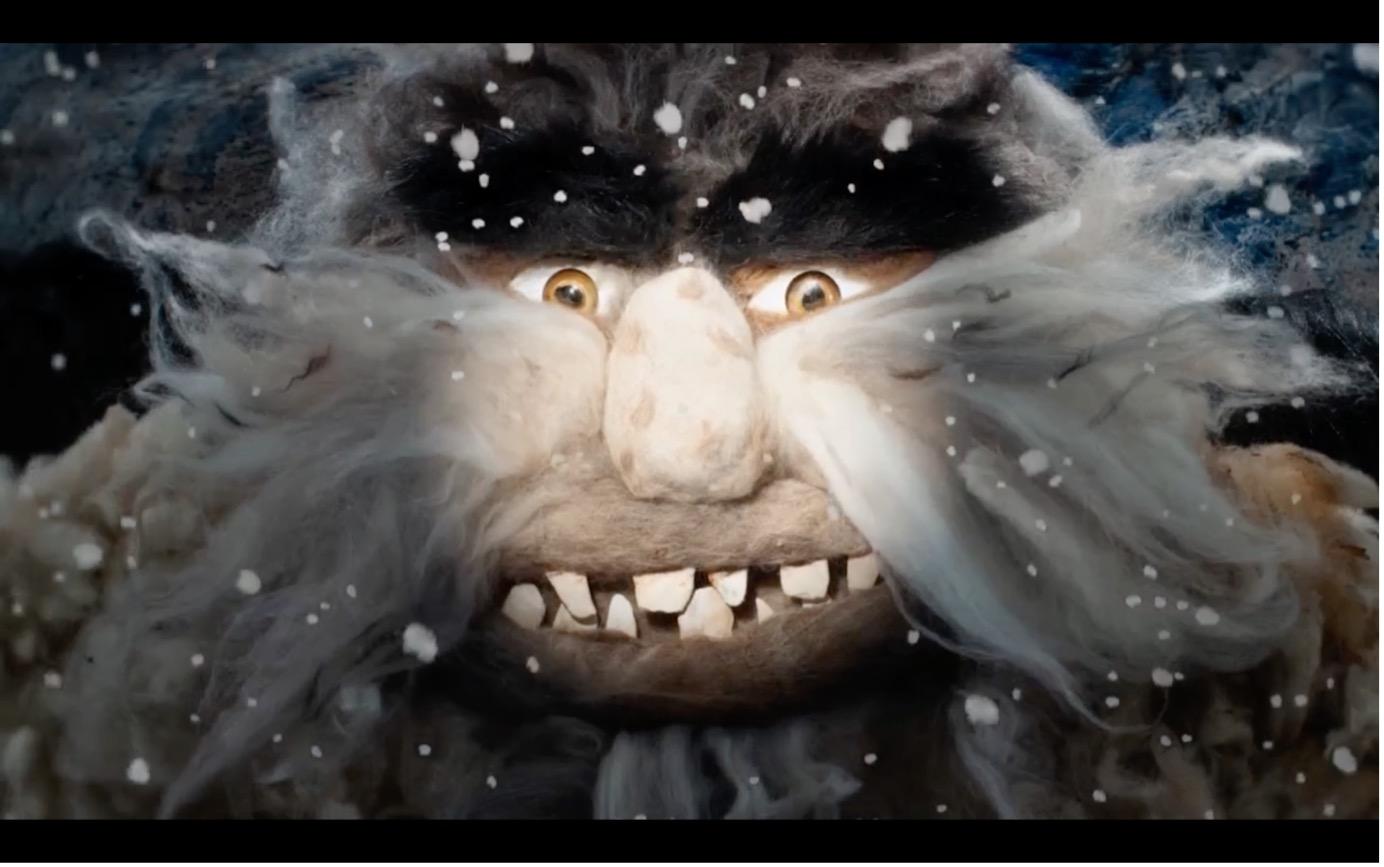 Das Bild zeigt das Gesicht einer trollartigen Kreatur im Schneesturm. Die Figur besitzt buschige schwarze Augenbrauen, große gelbliche Augen, eine breite Nase und einen wilden weißen Bart, der fast das gesamte Gesicht umrahmt. Zwischen den zerzausten Haaren blitzen unregelmäßige, leicht schiefe Zähne hervor. Schneeflocken treiben dicht vor dem Gesicht vorbei und verstärken die raue, winterliche Atmosphäre.