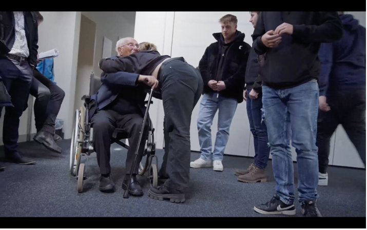 Ein älterer Mann im Rollstuhl wird von einer jüngeren Person umarmt, während mehrere Jugendliche die Begegnung beobachten. Die Aufnahme vermittelt durch die herzliche Geste und die Nähe der Anwesenden eine Atmosphäre von Verbundenheit und Anteilnahme.