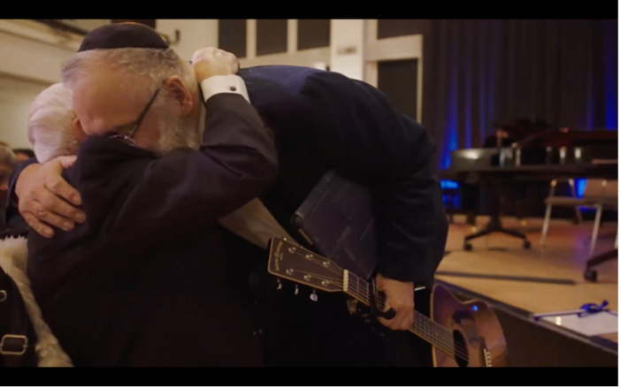 Ein älterer Mann umarmt einen bärtigen Musiker, der eine Gitarre in der Hand hält und sich zu ihm hinunterbeugt. Die innige Umarmung vermittelt große Herzlichkeit, Nähe und emotionale Verbundenheit.