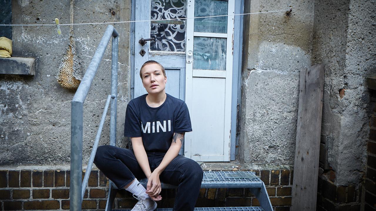 Susanne Heinrich, Regisseurin von Das melancholische Mädchen: sie sitzt vor einer Haustür auf einer Metalltreppe. Sie trägt eine schwarze Hose und ein schwrzes T-Shirt auf dem es "Mini" in weiße Schrift zu lesen ist. Sie guckt in die Kamera. 