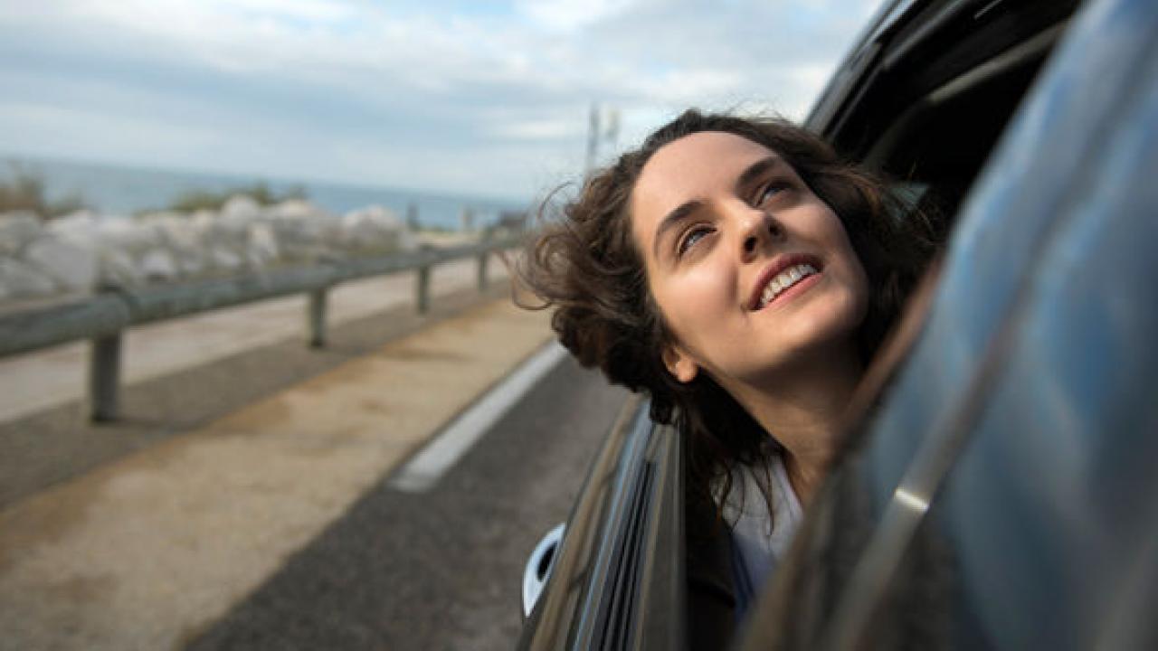 Eine junge Frau mit dunklen Haaren guckt lächeln aus dem Fenster eines Autos aus. Im Hintergrund ist die Straße und das Meer. 