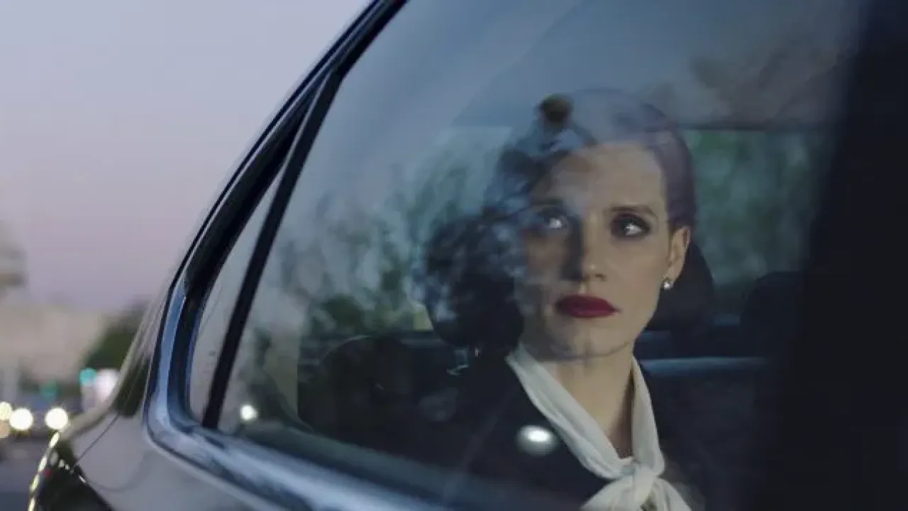 Eine Frau mit gebundene Haare, eleganter Kleidung und roten Lippenstift sitzt im Hintersitz eines schwarzen Autos und guckt ernst aus dem Fenster raus.