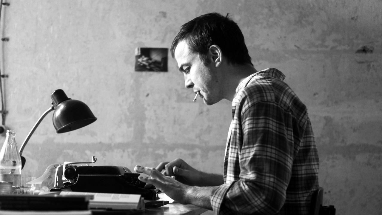 Szenenbild aus dem Drama "Lieber Thomas": Ein junger Mann sitzt an einem Tisch und tippt auf der Schreibmaschine. Er hat eine Zugarette im Mund.